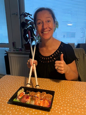 En kvinna gör tummen upp och har två vita käppar i höger hand likt ätpinnar. På bordet framför henne står en låda med sushi 