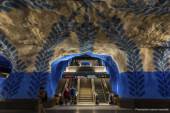 En tunnelbanenedgång där två rulltrappor finns centrerade på bilden, emellan dem finns en trappa. En man och kvinna kliver av den vänstra rulltrappan. Tunnelbanestationens väggar är blå- och vitmålade med mönster i en mörkare blå nyans.