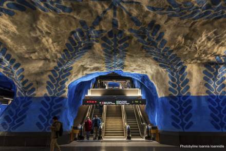 En tunnelbanenedgång där två rulltrappor finns centrerade på bilden, emellan dem finns en trappa. En man och kvinna kliver av den vänstra rulltrappan. Tunnelbanestationens väggar är blå- och vitmålade med mönster i en mörkare blå nyans.